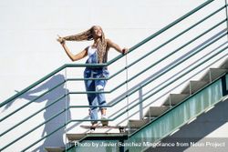 Happy female in denim overalls standing on skateboard on stairs playing with her hair 0JXlw4
