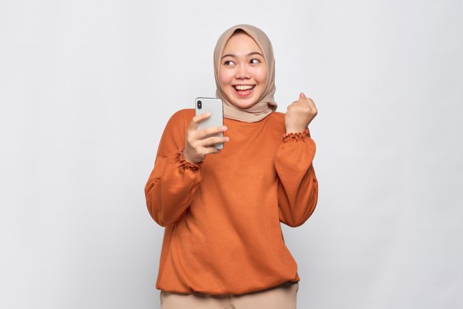 Smiling Muslim woman in headscarf and orange shirt with phone and celebratory fist