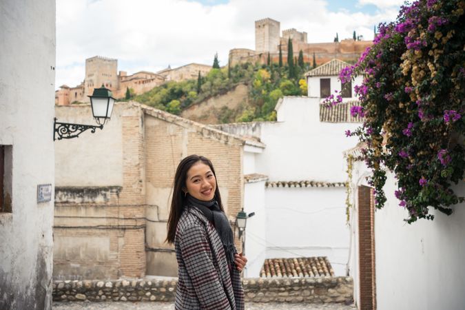 Asian female traveler in warm wear smiling among old buildings near big castle