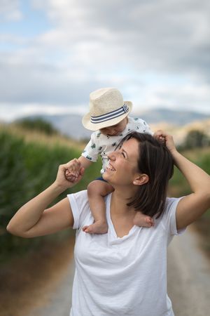 Adorable kid in hat on shoulders and looking at mother