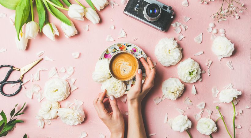 Coffee, camera, tulips artfully arranged on pink background, with hands on coffee cup