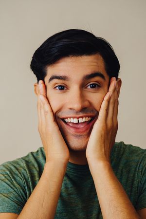 Portrait of Hispanic male in neutral room with hands on both cheeks, vertical