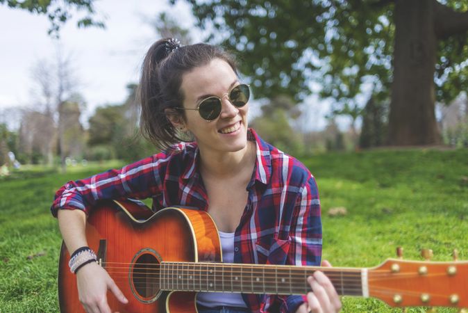 Front view of a young hipster woman wearing sunglasses, sitting on grass in a park while enjoying playing guitar