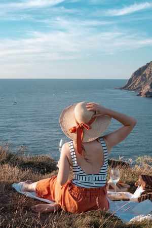 Woman with glass of drink on picnic mat looking at sea and mountains