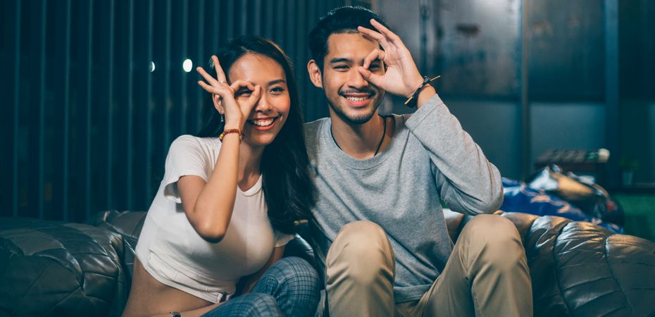 Asian young couple holding hand gesture on eye while sitting together and smiling