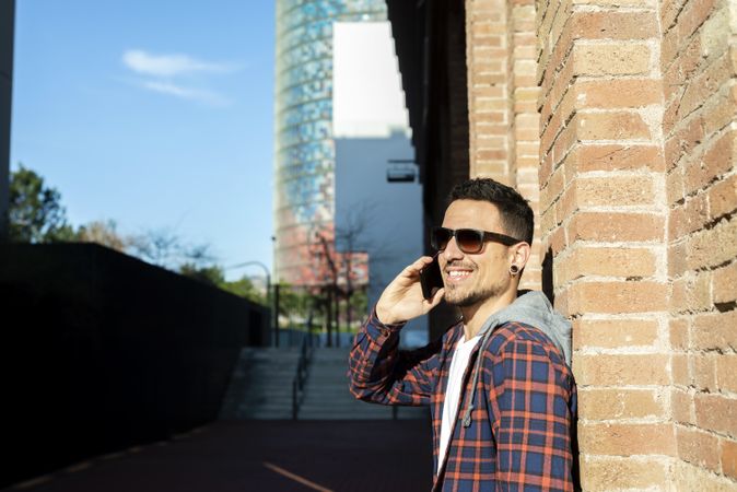 Happy man leaning on a brick wall wearing sunglasses while using a smartphone