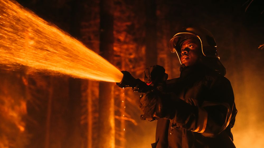 Black Fireman Holds High-Pressure Water Hose in Hands, Battling Bushfire.