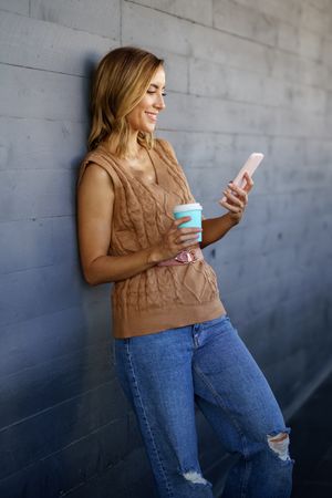 Cheerful female with smartphone and coffee leaning on wall
