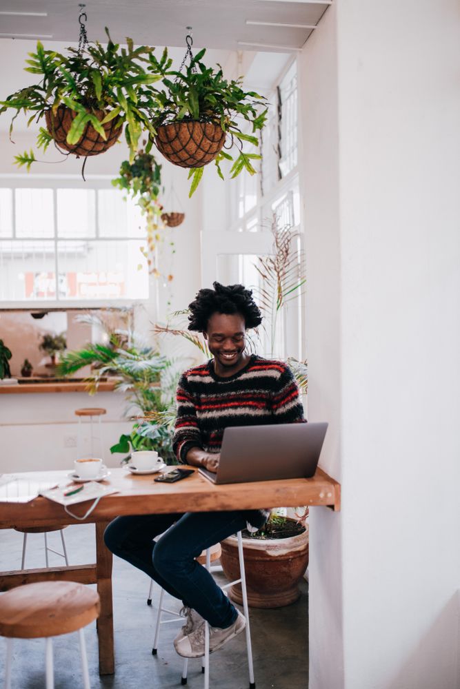 Man smiling while working on his computer at a coffee shop with hangplants