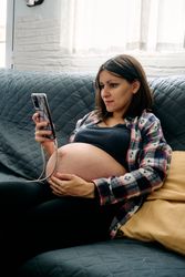 A young woman with a very advanced pregnancy is sitting on the couch looking at and talking on her cell phone.