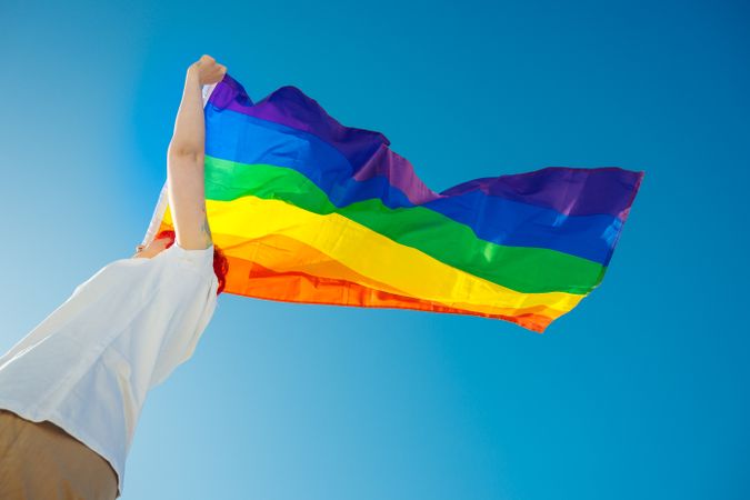 Low angle view woman waving rainbow flag under blue sky
