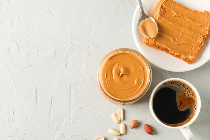 Glass jar with peanut butter, peanut, cup of coffee, spoon and peanut butter sandwich on grey background, top view and space for text