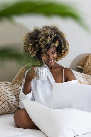 Black woman drinking coffee while sitting on bed with laptop