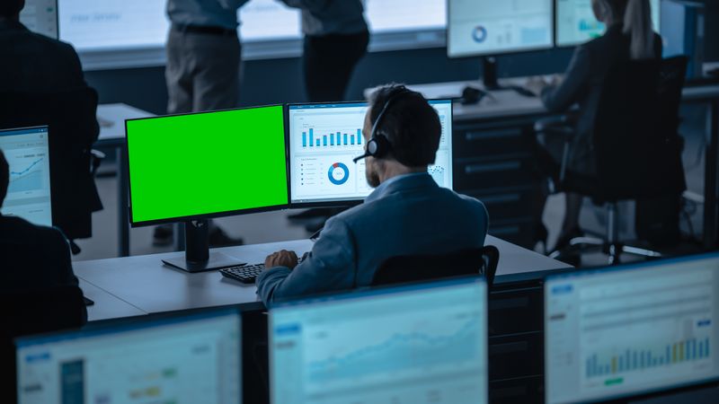 Data Analyst With Headset Examines Financial Charts on Dual Monitors.