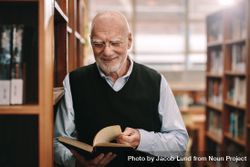 Cheerful older man standing in a library holding a book 4d1llb