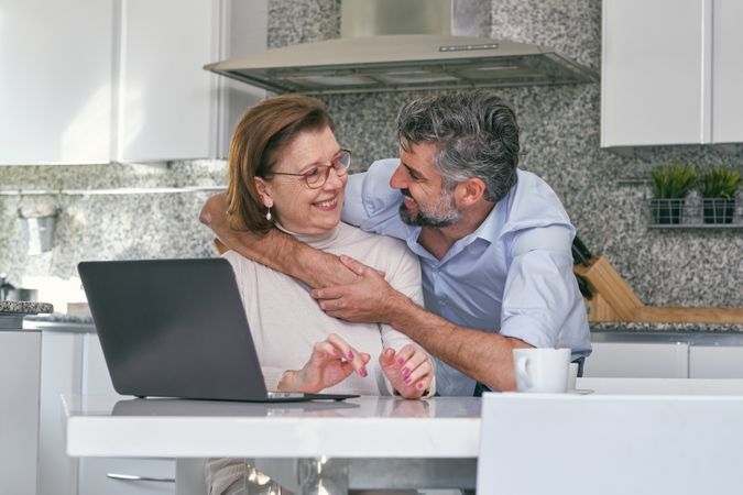 Adult son hugging senior mother at table with laptop