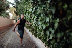 Mature man stretching leg holding botanical wall in street