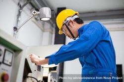 Side view of Asian man in blue jumpsuit and yellow hard hat working with machinery 0vNNL0