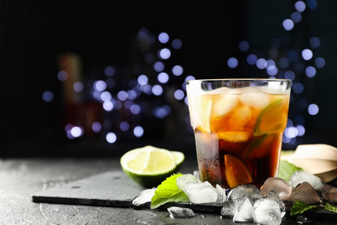 Glass with cola and lime slices, juicer, ice, mint against blurred lights background