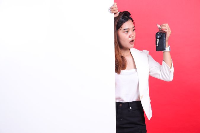 A woman with brown hair and a jacket is holding a car key and standing behind a poster with a surprised face, isolated on a red background