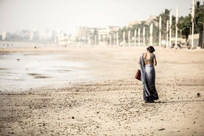 Woman in gray sari and holding a suitcase walking on brown sand