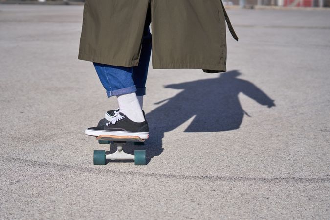Crop man in sneakers and raincoat riding skateboard