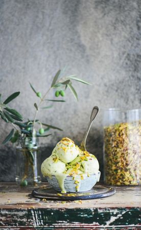 Bowl of melting pistachio ice cream with spoon and grey background with leaves and glass container