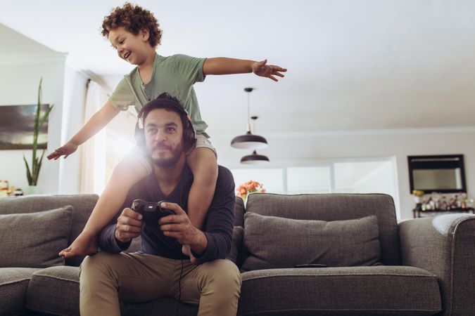 Man playing video game sitting on couch at home with his son sitting on his shoulders