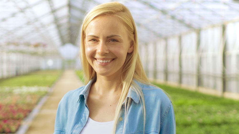 Shot of Beautiful Blonde Gardener Stands Smiling in a Greenhouse Full of Colorful Flowers.