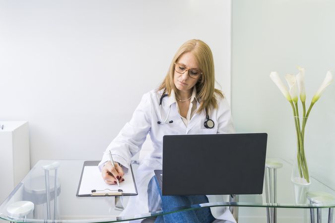 Female doctor working on laptop and writing on clipboard in medical office.