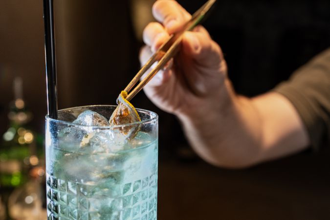 Bartender preparing gin and tonic cocktail with ice and lemon peel, closeup of glass and hand