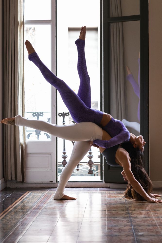 Flexible woman supporting female friend on back while doing Eka Pada Adho Mukha Svanasana yoga pose