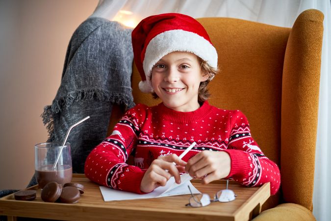 Cheerful boy in Santa hat drawing at table with chocolate