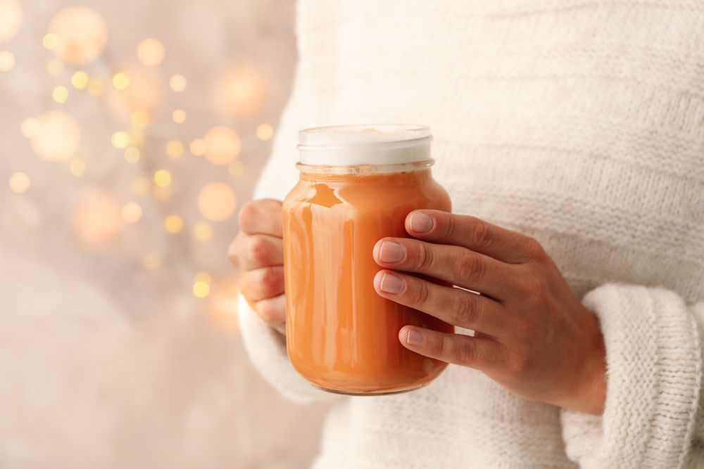 Woman hold glass jar of pumpkin latte against brown background with blurred lights