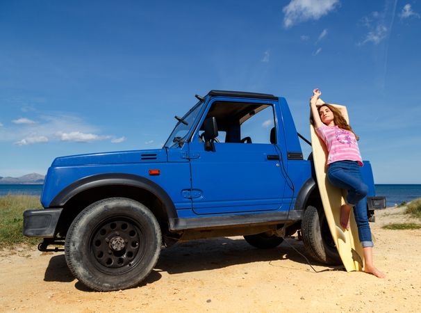 Woman leaning on surfboard near car at seaside