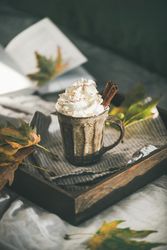 Mug of warm drink topped with whipped cream, on wooden trey with leaves, with book in background