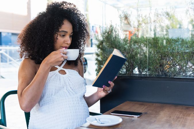 Pregnant Black woman reading book and drinking coffee