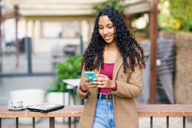 A young woman is sitting outdoors, enjoying a warm cup of coffee while using her smartphone