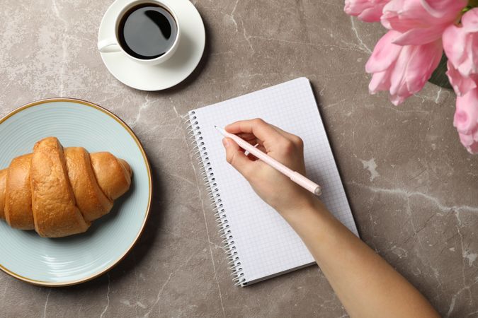 Female hand hold pen on grey background with notebook, coffee, tulips and croissant, top view