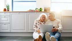 Mature couple sitting on the kitchen floor with their laptop and wine