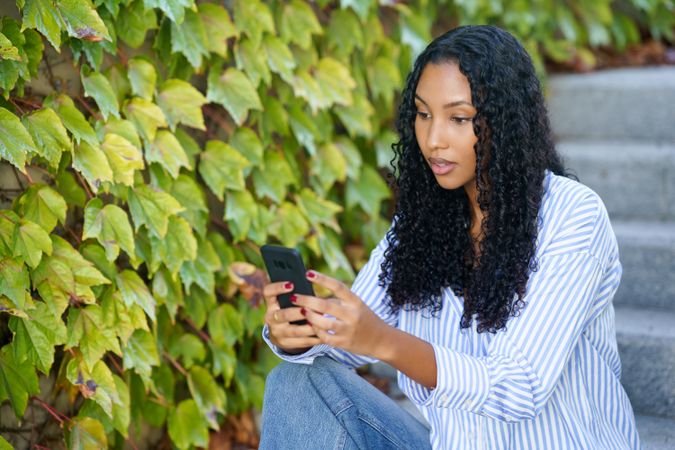A young woman using her smartphone outdoors, surrounded by greenery, enjoying the day