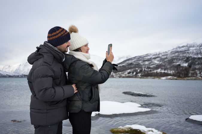 Young couple shooting with smartphone on pond beach against snowy hills in cold overcast weather