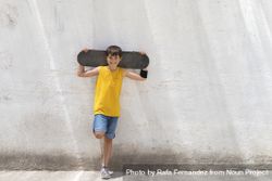 Young smiling boy leaning on wall holding a skateboard while looking camera on sunny day 5wXpGm