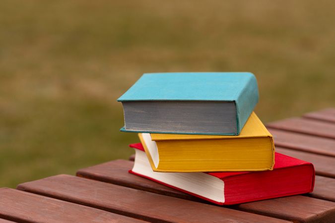 Colorful hardcover books stacked outdoors on wooden park bench.