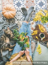 Woman with cat on blanket sitting on colorfully tiled balcony with fall leaves, squash, and mug 5QKXVb