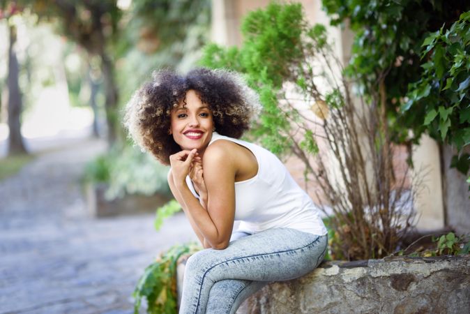 Young Black woman with afro hairstyle smiling while sitting on cement wall outside