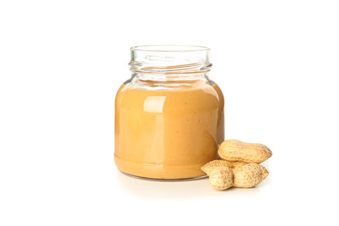Creamy peanut butter in glass jar and peanut isolated on light background. A traditional product of American cuisine