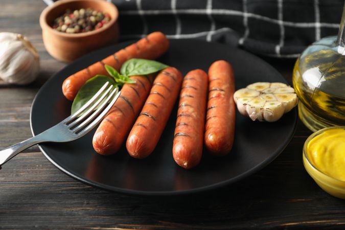 Grilled sausages or hot dogs on a fork on dark ceramic plate with garlic