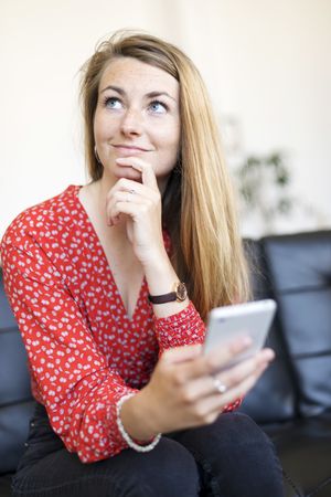 Beautiful woman using mobile phone and looking away thoughtfully while sitting on sofa at home
