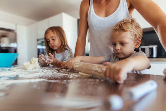 Little boy with his mother and sister preparing dough with rolling pin on kitchen table